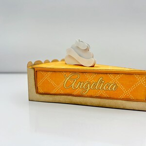 May include: A yellow and gold pie-shaped box with a white whipped cream design on top. The box has a decorative pattern and the name "Angelica" written on it.