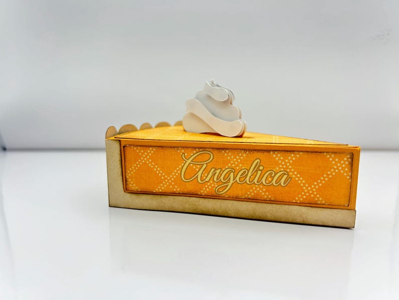 May include: A yellow and gold pie-shaped box with a white whipped cream design on top. The box has a decorative pattern and the name "Angelica" written on it.