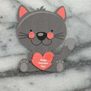 May include: A gray cat holding a red heart with the text "Happy Valentines Day!" printed on it.