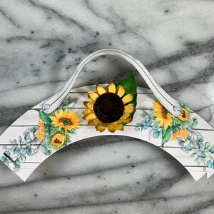 May include: White cupcake holder with a handle, featuring a sunflower design and a woodgrain pattern. The holder is decorated with sunflowers and eucalyptus leaves.