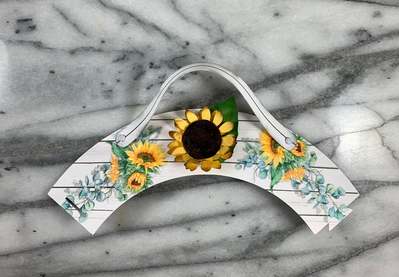 May include: White cupcake holder with a handle, featuring a sunflower design and a woodgrain pattern. The holder is decorated with sunflowers and eucalyptus leaves.