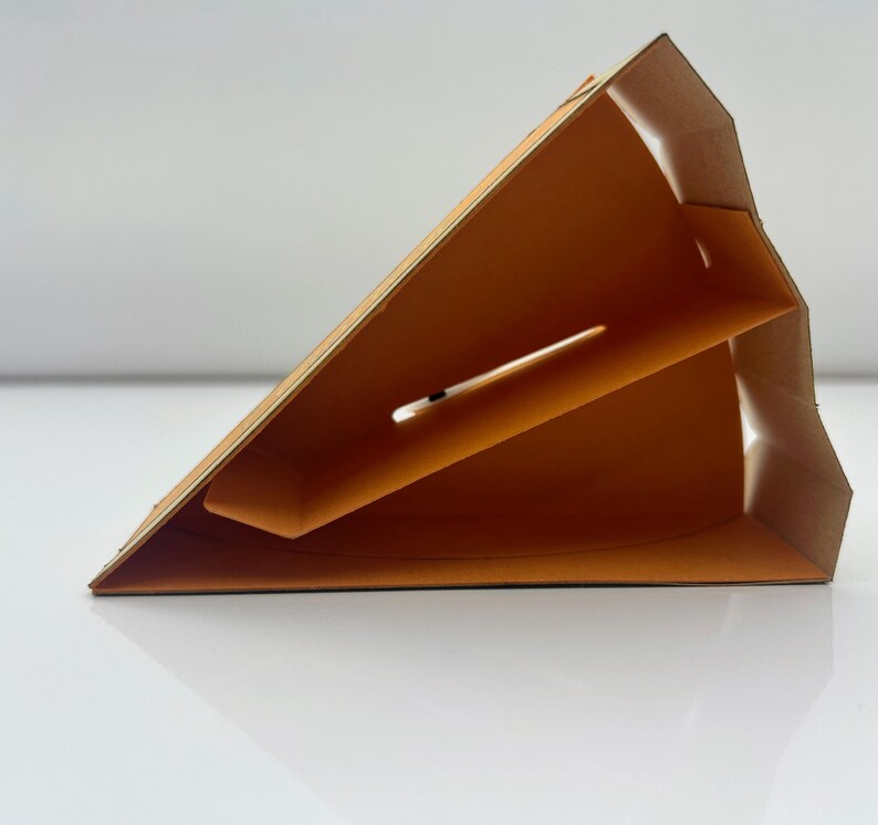 May include: A cardboard triangle-shaped box with a brown exterior and an orange interior. The box has a slot on the side and is designed to hold a single item.