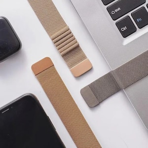 May include: Three metallic mesh watch bands in gold, rose gold, and dark gray tones. Each band features a flat rectangular magnetic clasp at the end. The bands are arranged near a black smartphone and a silver laptop keyboard with visible keys.