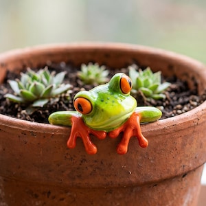 May include: A small, green glass frog figurine with large, bright orange eyes and orange feet, perched on the rim of a terracotta-colored clay flower pot containing dark soil and small, green succulent plants with pointed leaves.