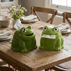 May include: Two glossy green ceramic frog-shaped tissue holders. Each frog features large white eyes with black pupils, pink cheek spots, and a wide mouth opening designed to dispense white paper tissues from the centre of the face.