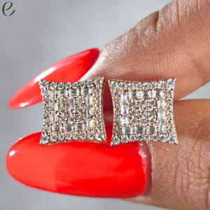 May include: A pair of square-shaped stud earrings featuring a central cluster of baguette-cut stones surrounded by a border of round brilliant-cut stones. The earrings are set in a metallic frame with a warm, rose-gold tone finish and clear stones.