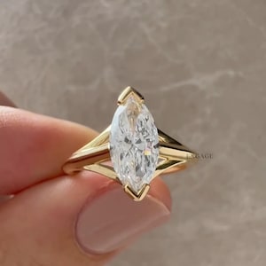 Könnte beinhalten: Ein Verlobungsring mit einem Diamanten im Marquiseschliff. Der Ring hat einen großen, klaren Diamanten in einem goldenen Band. Das Wort "ENGAGE" ist auf dem Ring zu sehen.