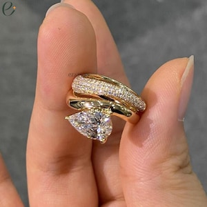 Könnte beinhalten: Ein goldfarbener Ring mit einem birnenförmigen, klaren Stein als Herzstück. Die Ringschiene ist in zwei Abschnitte unterteilt, wobei ein Teil mit kleinen klaren Steinen im Pavé-Stil besetzt ist und der andere eine glatte, polierte Oberfläche aufweist.
