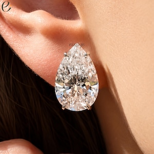 May include: A pear-shaped diamond stud earring featuring a clear, faceted stone set in a silver-toned metal prong setting. The word engage is printed in small black letters above the earring. The jewelry is positioned on an earlobe.