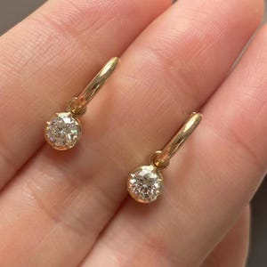 Puede incluir: Un par de pendientes de aro de oro, cada uno con una piedra preciosa transparente, redonda y facetada, suspendida en la parte inferior. Los aros son de oro pulido y las piedras están sujetas por pequeñas garras metálicas.