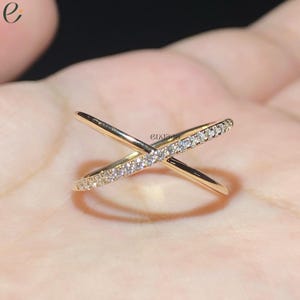 May include: A gold-toned crossover ring featuring an X-shaped design with one band encrusted with small clear stones and the other band featuring a smooth polished finish. The word engage is printed in small black letters on the ring surface.