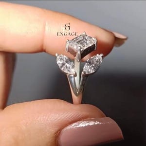 Può includere: Un anello d'argento con un diamante centrale taglio smeraldo e due diamanti taglio marquise. Il design dell'anello ricorda un fiore. L'anello è tenuto dalle dita di una persona. Il logo "ENGAGE" è visibile.