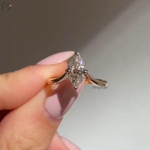 May include: A gold ring with a marquise-cut diamond center stone. The diamond is set with small accent stones. The ring has a delicate, curved band. The word "engage" is written on the side of the ring.
