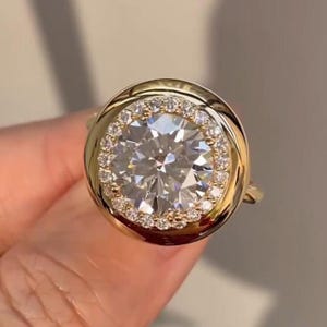 Op de afbeelding: Een gouden ring met een grote, ronde, heldere edelsteen omringd door een halo van kleinere stenen. De band en zetting van de ring zijn goudkleurig, wat een luxueuze en elegante uitstraling creëert. De centrale steen schittert helder.