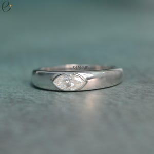 Può includere: Un anello d'argento con un diamante taglio marquise. Il diamante è incastonato in una lunetta e la fascia è liscia e lucida. La parola "engage" è visibile sull'anello.