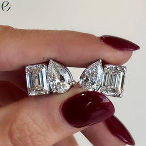 Peut inclure: Une paire de boucles d'oreilles composée d'une pierre taille émeraude et d'une pierre taille poire transparentes serties dans un métal argenté. Le mot engage est imprimé en petites lettres noires au-dessus des pierres sur la gauche.