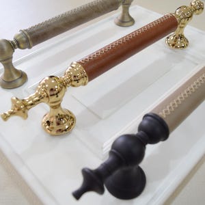 Puede incluir: Tres tiradores decorativos para armarios de diferentes estilos. Uno es de bronce con un agarre texturizado, otro tiene un agarre de cuero marrón con detalles dorados, y el tercero es negro con un agarre beige y costuras blancas.