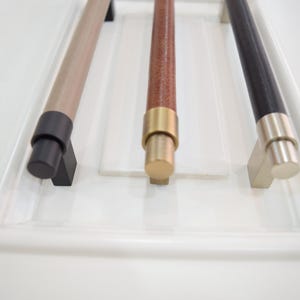 Peut inclure: Trois poignées de meubles présentées. La première est marron clair avec une base noire, la seconde est marron avec une base dorée et la troisième est noire avec une base argentée. Chaque poignée est cylindrique.