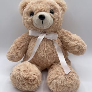 Puede incluir: Un oso de peluche marrón claro con una cinta blanca atada alrededor del cuello. El oso tiene una textura suave y esponjosa, ojos negros y una nariz negra cosida. El oso está sentado.
