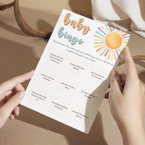 以下が含まれることがあります： カラフルな太陽のグラフィックが描かれたベビーシャワービンゴカード。カードは、16個の正方形のグリッドになっており、さまざまなビンゴプロンプトが書かれています。たとえば、「あなたと同じ月に生まれた人」や「青い車に乗っている人」などです。