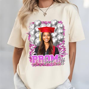 Pu&ograve; includere: T-shirt color crema con un design grafico per un saluto al ballo di fine anno. Il design include una foto di una persona sorridente con cappello e toga di laurea, circondata da palloncini argentati, glitter rosa e le parole "PROM Send Off."