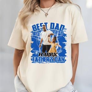 K&ouml;nnte beinhalten: Cremefarbenes T-Shirt mit blauem Aufdruck "BEST DAD EVER" und "HAPPY FATHER'S DAY". Die Grafik zeigt ein Foto von Vater und Kind. Das T-Shirt hat einen l&auml;ssigen Stil.