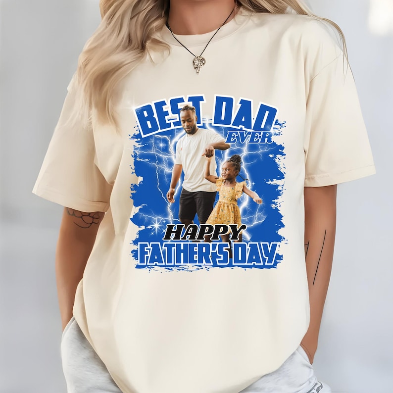 K&ouml;nnte beinhalten: Cremefarbenes T-Shirt mit blauem Aufdruck "BEST DAD EVER" und "HAPPY FATHER'S DAY". Die Grafik zeigt ein Foto von Vater und Kind. Das T-Shirt hat einen l&auml;ssigen Stil.