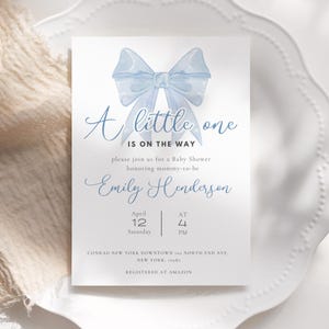 May include: A baby shower invitation with a blue watercolor bow and the text "A little one is on the way". The invitation is for Emily Henderson and is scheduled for April 12th at 4 PM. The event will be held at Conrad New York Downtown, 102 North End Ave, New York, 10282. The invitation states that the guest of honor is registered at Amazon.