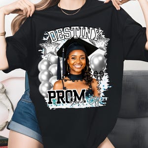 Pu&ograve; includere: T-shirt nero con un design a tema laurea. Il design include il testo "DESTINY PROM Send Off" con una foto di una persona sorridente con cappello e toga di laurea, circondata da palloncini e stelle argentate.