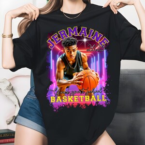May include: Black t-shirt featuring a basketball player holding a basketball. The shirt has the name "Jermaine" and the year "2023" in a purple and yellow design. The text "Your sport school BASKETBALL" is also visible.