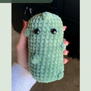 May include: A light green, plush, crocheted dinosaur-shaped toy with black button eyes. The toy has small, rounded ears and tiny pink claws. The text "Crochet Pattern" is in a green rectangle in the upper left corner.