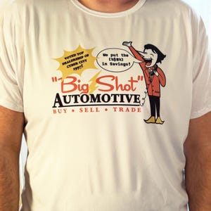 May include: White t-shirt featuring a cartoon advertisement for "Big Shot Automotive." The design includes a smiling salesman, text that reads "Voted Top Dealership of Cyber City 1997!" and the slogan "We put the [%$#%] in Savings!"