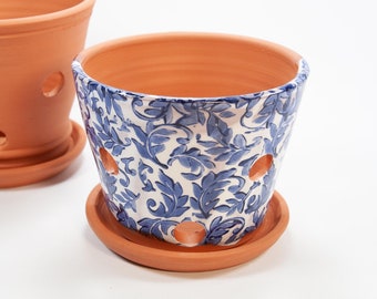 May include: A white ceramic planter with a blue floral pattern and drainage holes. The planter has a matching terracotta saucer.