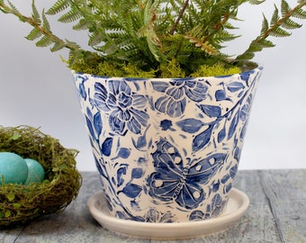 May include: A white ceramic planter with a blue floral and butterfly design. The planter is filled with a green fern plant.