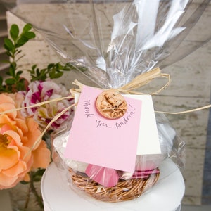 K&ouml;nnte beinhalten: Eine rosa und wei&szlig;e Geschenkbox mit einem braunen Weidenkorb im Inneren. Die Box ist in Klarsichtfolie verpackt und mit einem braunen Band gebunden. Ein kleines Keramik-Etikett mit einem Vogelmotiv ist am Band befestigt. Das Etikett lautet "Danke Andria!"