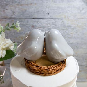 May include: Two white ceramic birds kissing on a brown woven nest cake topper.