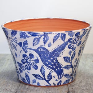 May include: A white ceramic planter with a blue and white floral pattern and a hummingbird design. The planter has a wide, round opening and a slightly flared rim.