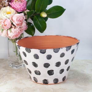 May include: A white ceramic planter with a black polka dot pattern. The planter has a wide, rounded base and a slightly flared rim. The planter is empty.