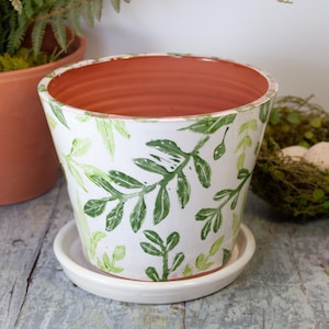 May include: White ceramic plant pot with a green leaf pattern, sitting on a matching saucer. The interior of the pot is terracotta. A smaller terracotta pot is visible in the background.
