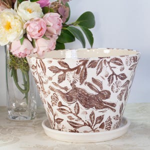 May include: White ceramic planter with a brown rabbit and floral design. The planter has a matching saucer.