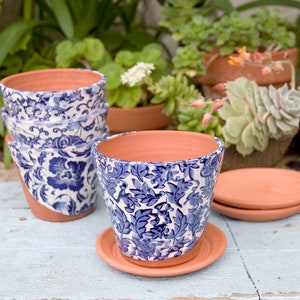 May include: Set of four blue and white patterned terracotta flower pots with a blue and white floral design. The pots are stacked on top of each other and are sitting on a light blue wooden surface.