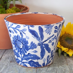 May include: A white ceramic flower pot with a blue floral design featuring dragonflies and flowers. The pot has a terracotta interior and is suitable for planting flowers or herbs. The pot is approximately 7 inches in diameter.