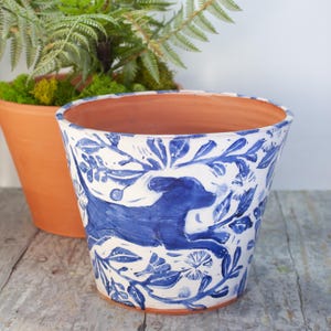 May include: A white ceramic planter with a blue and white design featuring a dog running through a floral pattern. The planter is a wide, rounded shape.