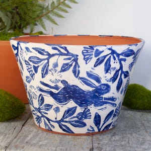 May include: White ceramic planter with a blue rabbit and floral pattern. The planter has a wide opening and a terracotta-colored interior. The design features a running rabbit and botanical elements, creating a nature-inspired aesthetic.