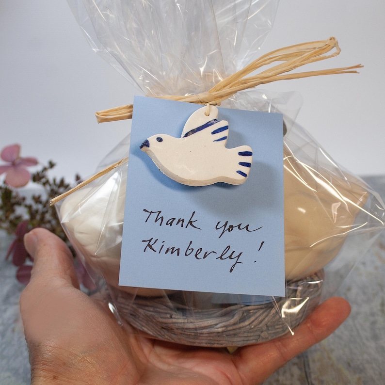 May include: A white ceramic dove with blue accents is attached to a blue gift tag with the handwritten message "Thank you Kimberly!" The dove is sitting on a nest of blue and white twine, wrapped in clear cellophane.