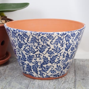 May include: A white ceramic planter with a blue and white floral pattern. The planter is wide at the top and tapers down to a narrow base.
