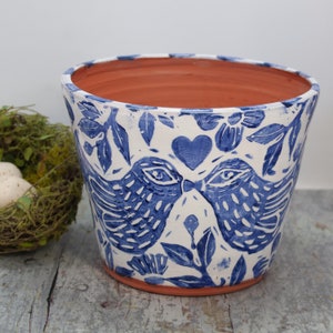 May include: A white ceramic planter with a blue and white floral design featuring two birds kissing. The planter has a terracotta rim.