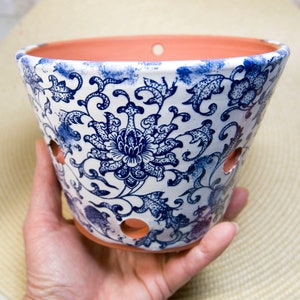 May include: A white ceramic hanging planter with a blue floral design. The planter has drainage holes and a rope for hanging.