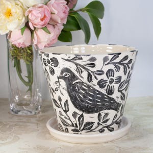 May include: A white ceramic planter with a black and white floral design featuring a bird. The planter has a matching saucer.