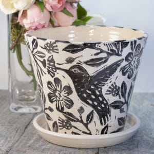 May include: A white ceramic planter with a black and white hummingbird and floral design. The planter has a matching saucer.